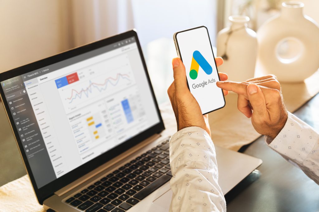 Man using a mobile phone with Google Ads on the screen and in the background a computer showing ad statistics.